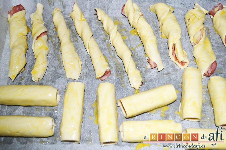 Aperitivo de hojaldre con salchichas o bacon, enrollar los paquetitos, ponerlos en una bandeja de horno sobre papel vegetal y pintar de huevo